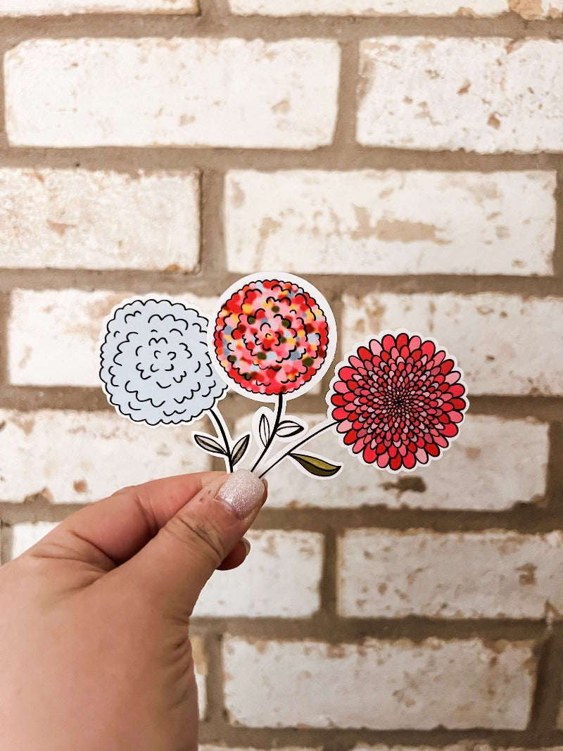 Three colorful floral stickers held in a hand against a white brick background. Stickers feature a blue flower, a multi-colored flower with pink, blue, and yellow petals, and a pink flower with a brown center. These stickers add a touch of whimsy and charm to any surface or project.