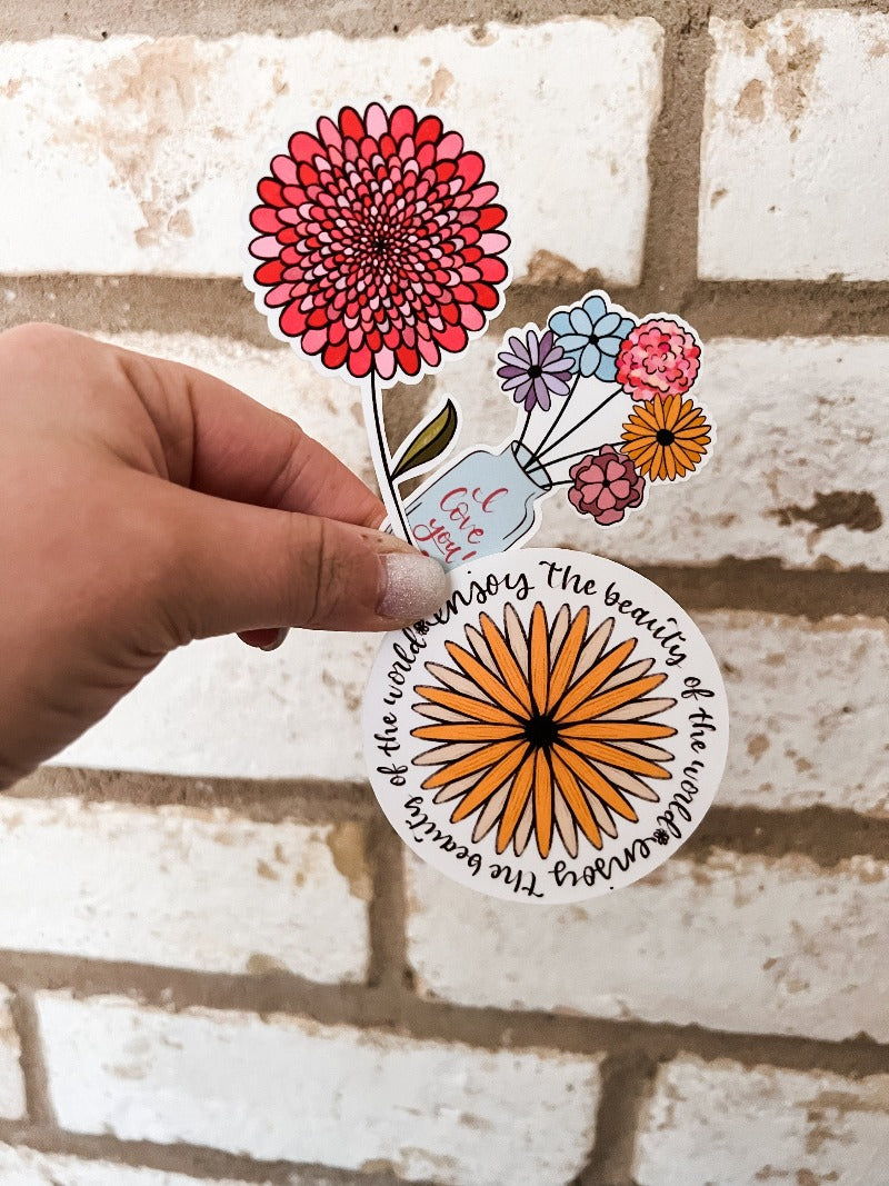 Three spring stickers in a hand over white brick: pink flower with brown center, colorful vase of flowers, and yellow chrysanthemum with 'Enjoy the Beauty of the World' message. Adds pop of color and positivity.