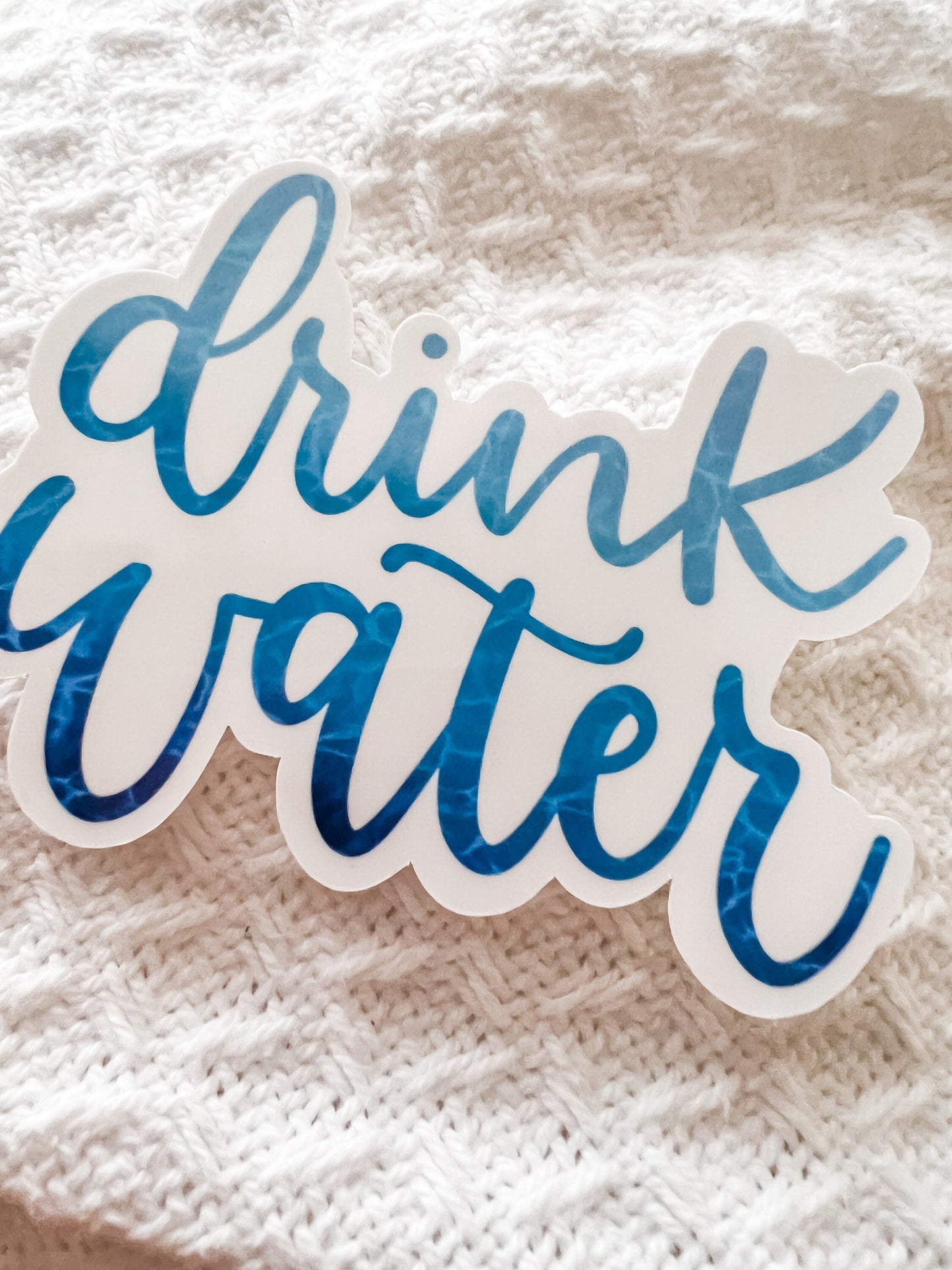 Close-up photo of a 'Drink Water' sticker featuring blue script lettering and a light blue water pattern on the letters. The sticker is designed for use on water bottles or laptops and is made from high-quality sticker paper.