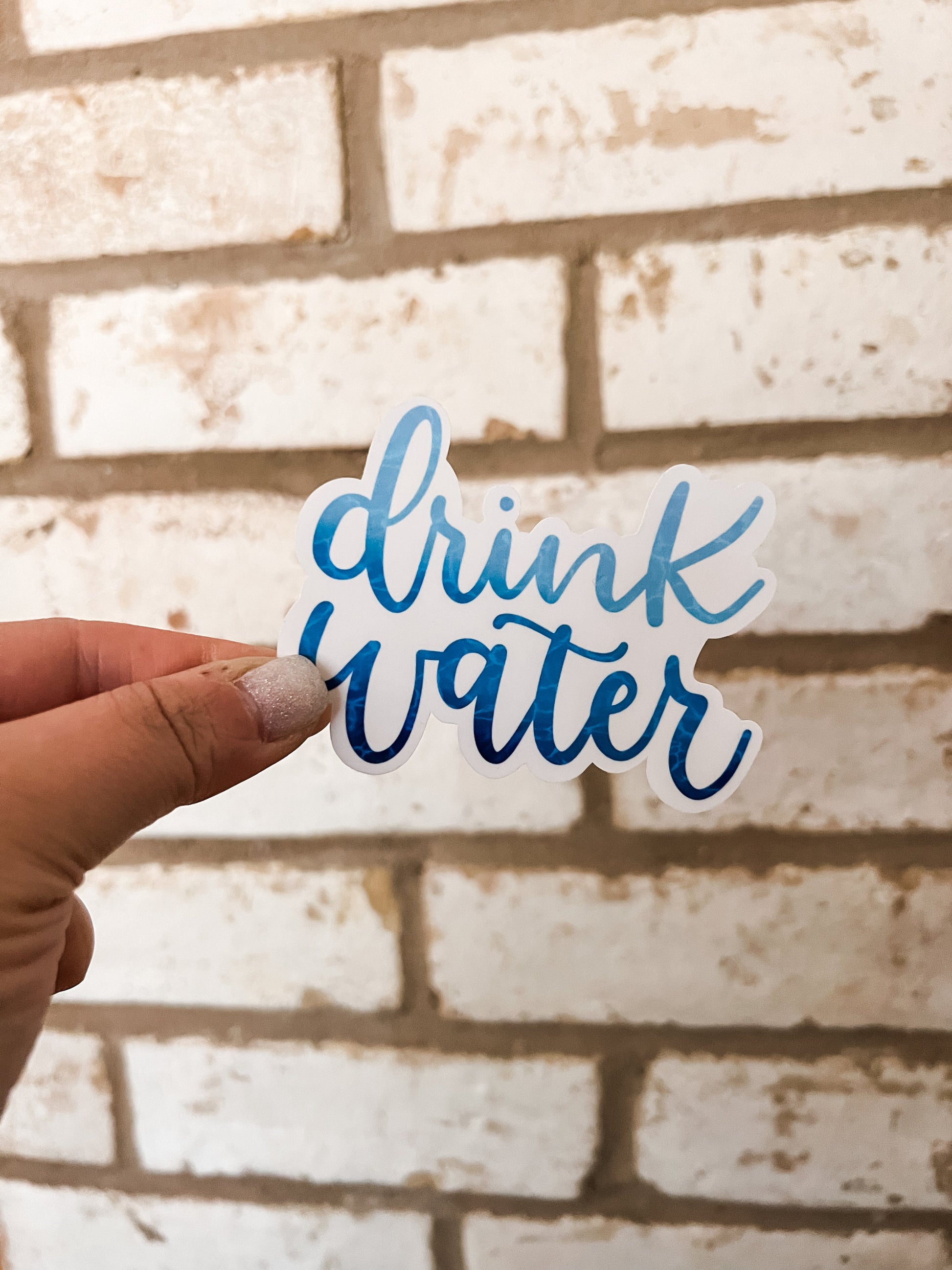 Close-up photo of a 'Drink Water' sticker featuring blue script lettering and a light blue water pattern on the letters. The sticker is designed for use on water bottles or laptops and is made from high-quality sticker paper.