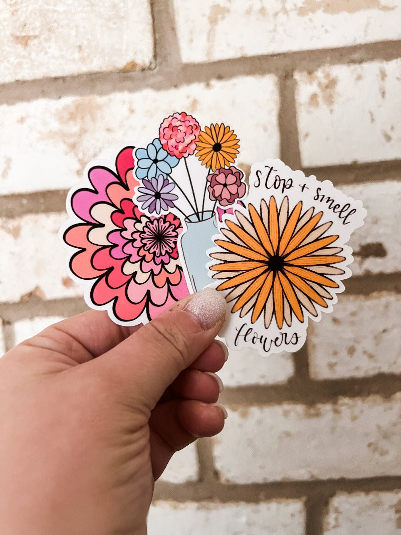 Three floral stickers held in a hand against a white background. Stickers feature a retro pink, yellow, and red flower with a black center, a vase of flowers in pink, yellow, and blue hues, and a yellow chrysanthemum with a brown center and the message 'Stop and Smell the Flowers'. These stickers are perfect for adding a cheerful and nostalgic vibe to any project or surface.